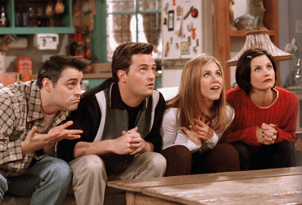 Foto com quatro dos seis protagonistas de Friends; em ordem da esquerda para direita sentados no sofá estão Joey, Chandler, Rachel e Monica olhando para frente inclinados com uma postura e expressão de expectativa, como se fossem ouvir uma história ou um segredo