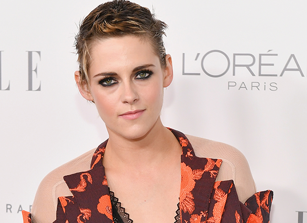 kristen-stewart-cabelo-curto-1 Kristen Stewart posando na frente de parede da loreal com vestido marrom e laranja e expressão levemente sorridente.