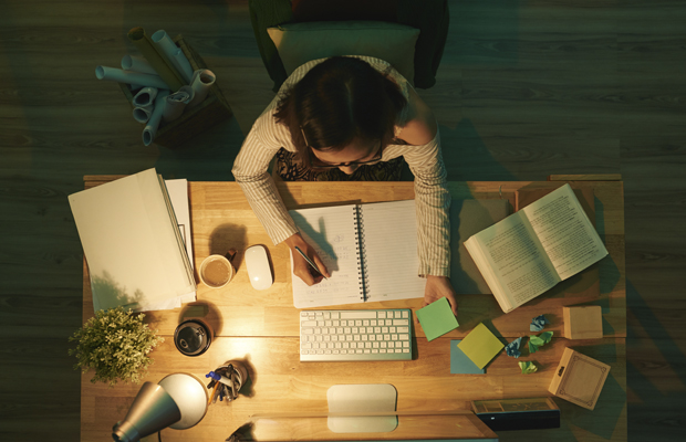 A imagem mostra uma mesa de estudos, vista de cima, com livros abertos, um teclado de computador e um mouse, post its coloridos, folhas de papel e uma luminária. Uma menina de cabelos compridos e escuros está sentada em frente a mesa, olhando para frente e escrevendo em um caderno.