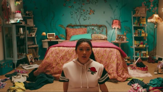 quarto-lara-jean-para-todos-os-garotos-que-ja-amei1 Quarto da Lara Jean de Para Todos os Garotos Que Já Amei; ela está no centro da cena com expressão assustada sentada no chão na frente da cama; a parede é decorada com flores e algumas roupas estão espalhadas pelo chão; ela usa uma blusa rosa com uma rosa bordada do lado esquerdo superior próxima ao coração; a roupa de cama é rosa com uma almofada verde turquesa; a parede é também verde turquesa com detalhes cor-de-rosa