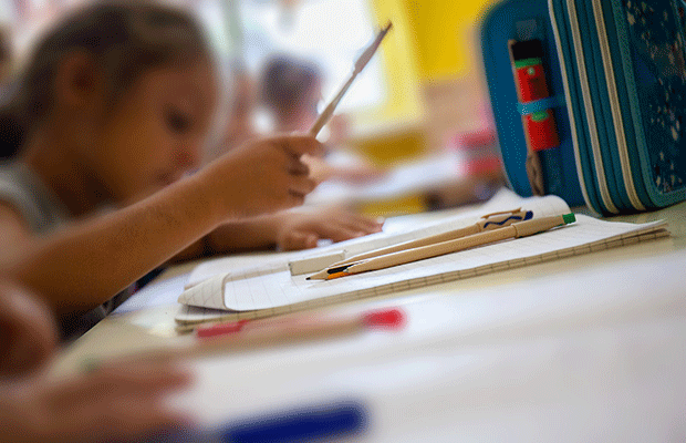 A imagem mostra uma mesa de estudos, com um caderno, lápis e estojo apoiados nela. Ao fundo da imagem, um pouco desfocada, é possível ver uma menina estudando.