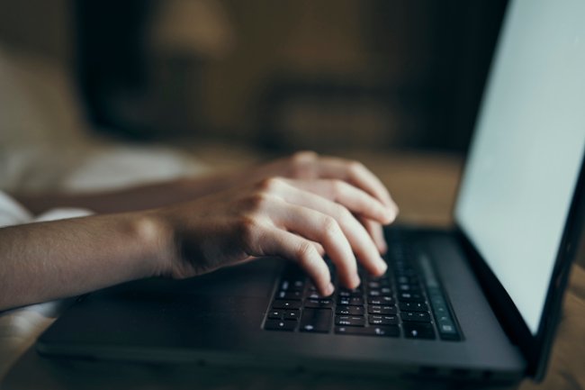 Inep divulga notas do Enem 2018: saiba como conferir! A imagem mostra duas mãos de uma pessoa branca digitando em um computador preto, com a tela ligada. O fundo da imagem é desfocado.
