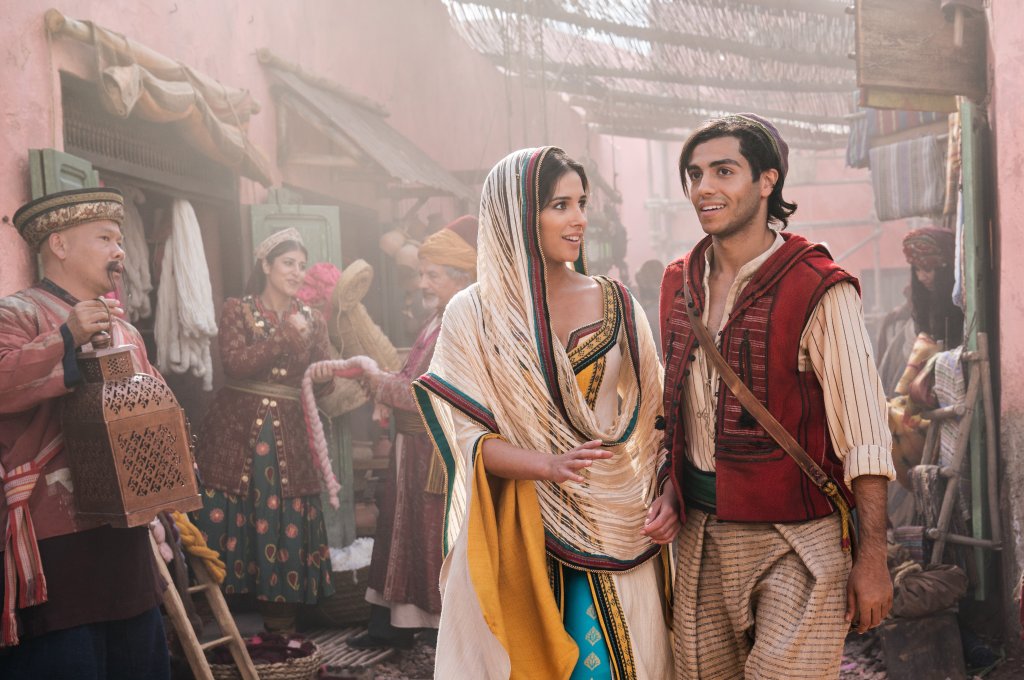 Naomi Scott e Mena Massoud são Jasmine e Aladdin no live-action da animação da Disney; na foto eles estão caracterizados como os personagens e andando na rua; Jasmine olha para Aladdin com expressão curiosa e ele está olhando pra frente e sorrindo