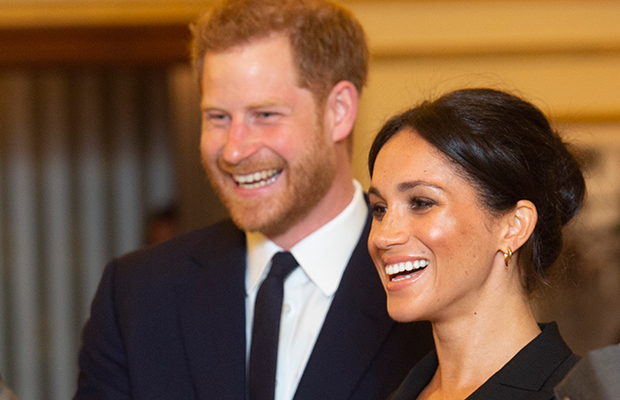 Príncipe Harry e Meghan Markle de perfil; os dois estão sorrindo observando algo; Harry usa um terno e gravata preta com camisa branca e Meghan usa roupa preta com cabelo preso