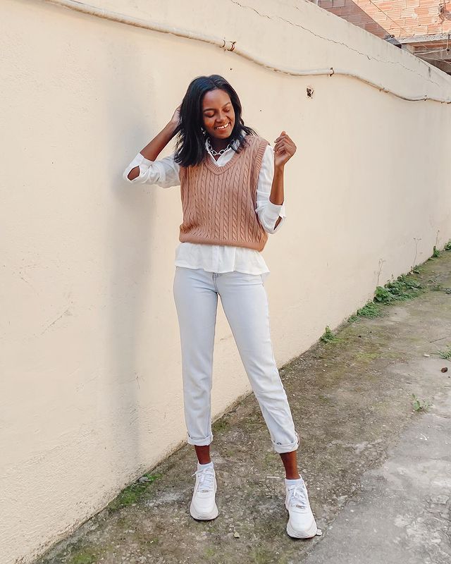 Garota usando colete de tricô marrom por cima de camisa branca, com calça jeans clara e tênis branco. Ela está sorrindo, com uma das mãos atrás do cabelo e o outro braço dobrado para cima.