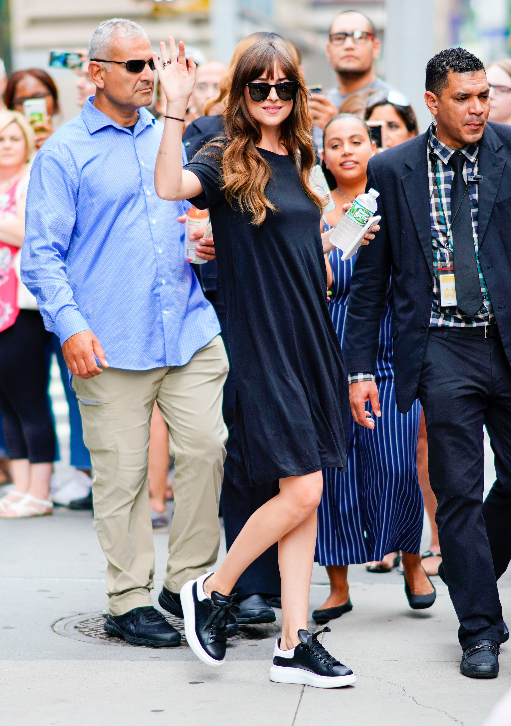 Dakota Johnson usando vestido preto, tênis preto e branco, óculos de sol e acenando para a foto