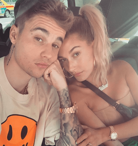 Selfie de Justin e Hailey Bieber; ele usa boné e uma camiseta bege; Hailey está de cabelo preto e roupa bege; eles estão no carro com as cabeças encostadas e Justin está apoiando o queixo em sua mão