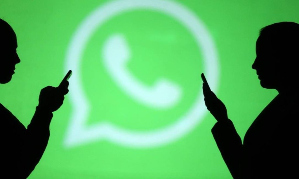 Duas pessoas seguram celulares e ao fundo está parede com o logo do Whatsapp