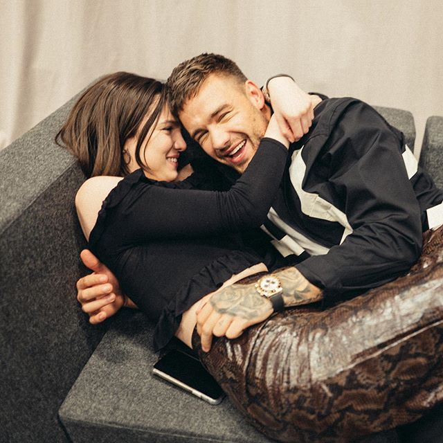 Liam Payne e Maya Henry deitados no sofá sorrindo; ela está abraçando o cantor pelo pescoço e encostando seu rosto no dele enquanto ele está rindo por cima dela