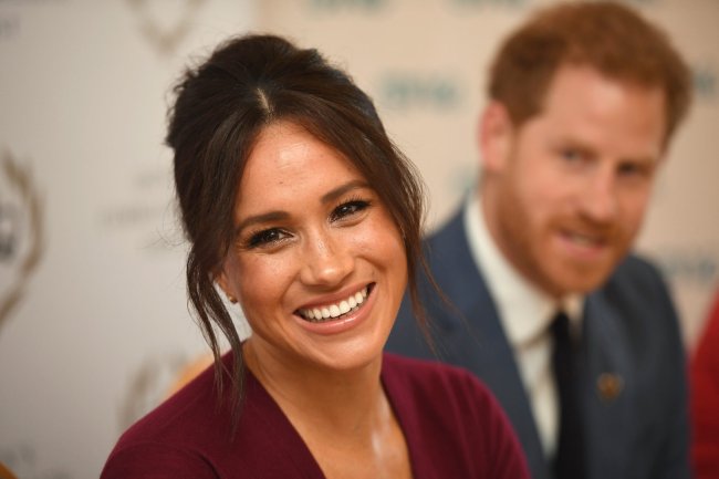 Meghan Markle sorrindo para foto; ela está com o cabelo preso e a franja divida ao meio com pontas soltas; ela usa uma roupa vinho