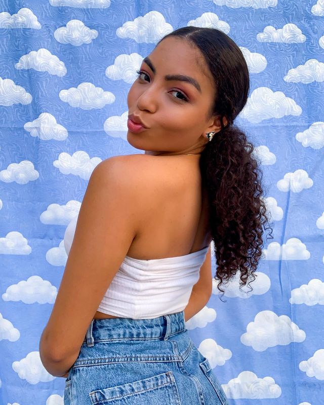 Foto da cantora Any Gabrielly em frente a uma parede azul com detalhes de nuvem. Ela usa um top cropped branco, shorts jeans, colar prata, brincos pratas, maquiagem natural com esfumado marrom e cabelo preso em com rabo de cavalo baixo. Ela olha para a câmera e faz biquinho.