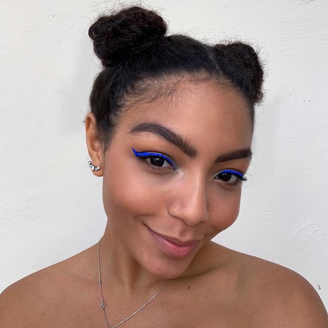 Selfie da cantora Any Gabrielly com foco no rosto e penteado. Ela usa uma maquiagem natural com delineado gatinho azul e cabelo preso no penteado space buns (dois coques altos). Ela olha para a câmera e sorri levemente.