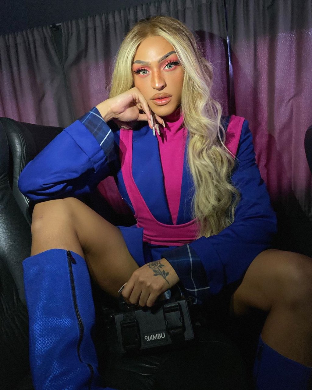 Pabllo Vittar posando para foto com a mão apoiada no rosto; ela está com expressão séria e usa uma roupa azul e rosa com bota de cano alto e cabelo solto