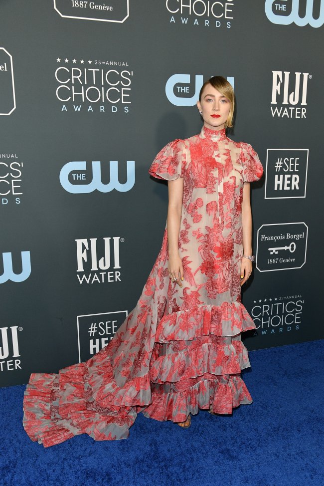 look-do-dia-saoirse-ronan Saoirse Ronan com vestido todo romântico com detalhes vermelhos e babados; ela está com expressão neutra, cabelo preso e usa batom vermelho posando no carpet do Critics Choice Awards promovido pela CW e pela FIJI que aparecem com seus logos no backdrop ao fundo da imagem