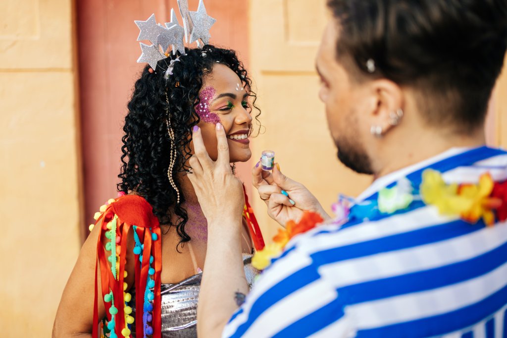 Foto no bloquinho de Carnaval de uma pessoa retocando o glitter de uma mulher que veste tiara, ombreiras e top prateado