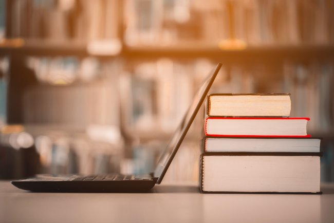 A imagem mostra um computador virado de lado em uma mesa e apoiado em uma pilha de quatro livros. Ao fundo da imagem, levemente desfocada, é possível ver uma estante com vários livros.