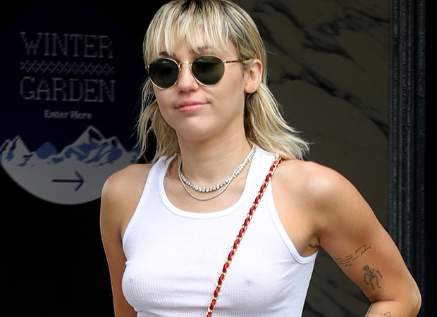 miley-cyrus-cabelo-mullet Miley cyrus com óculos escuro, regata branca, óculos preto, cabelo loiro e expressão sorridente.