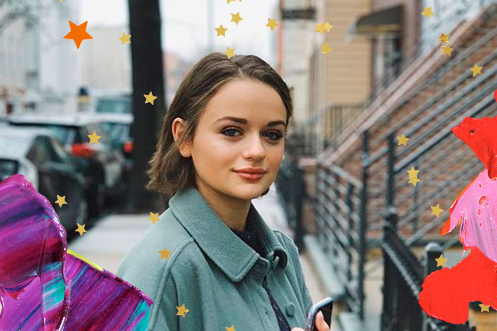 Joey King usando jaqueta azul, com o corpo de lado e olhando com expressão facial séria para a câmera, mas um leve sorriso de canto de boca. Na montagem, há estrelinhas douradas e laranjas por cima da foto, assim como tintas em roxo, laranja e rosa.