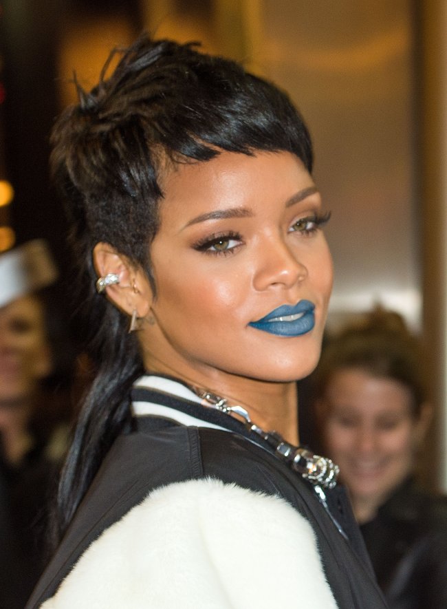 Rihanna posando com batom azul, olhando para o lado sorridente, com camisa preta e branca.