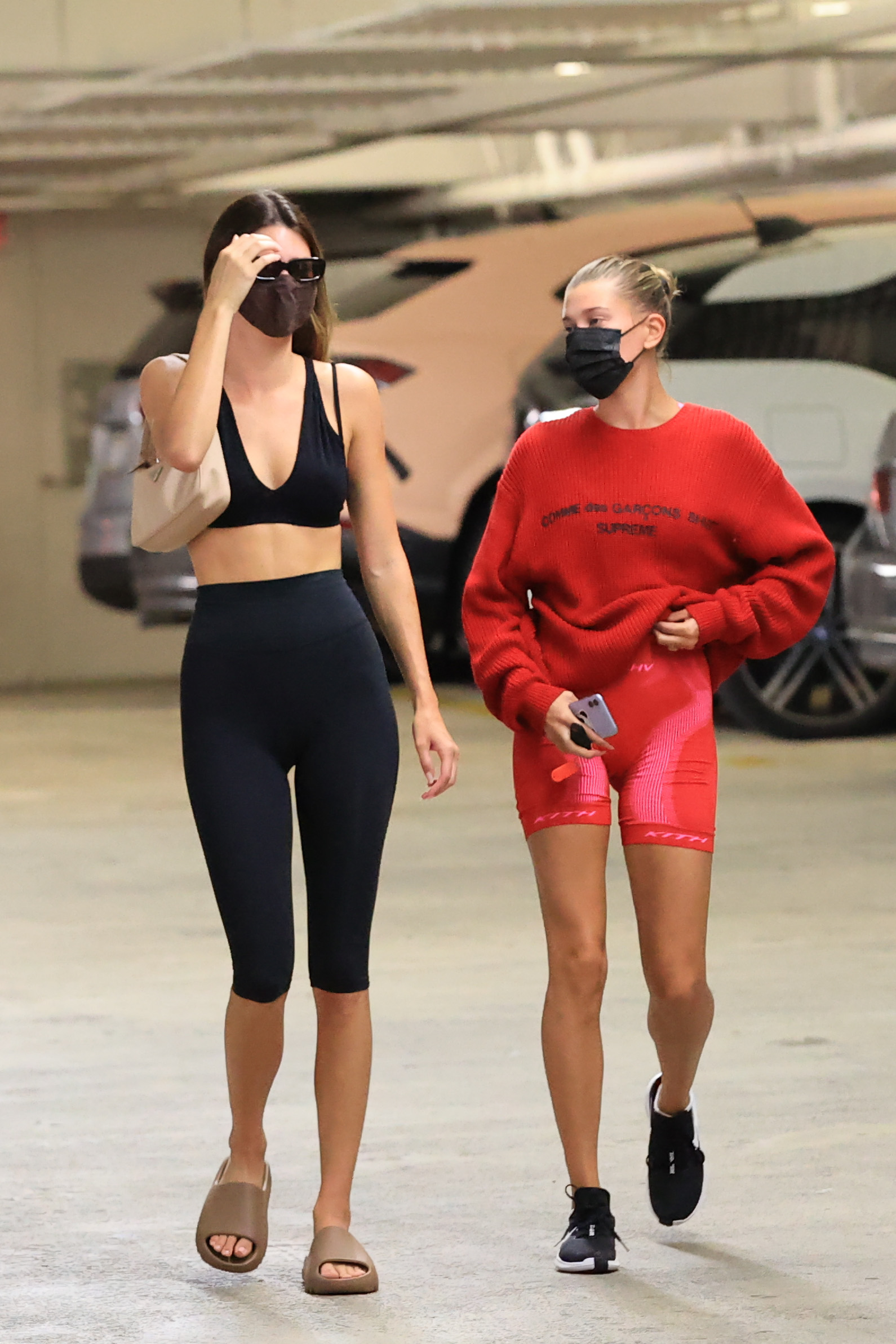 look-do-dia-kendall-jenner-biker-shorts Kendall Jenner usando look preto formado por top de academia e biker shorts com chinelo slide e shoulder bag, e Hailey Bieber usando look vermelho formado por moletom, biker shorts e tênis