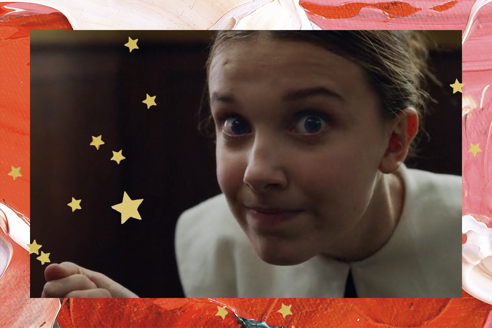 Foto de Millie Bobby Brown em cena de Enola Holmes; a atriz está com os olhos arregalados e pressionando os lábios sorrindo levemente com o cabelo preso e repartido ao meio; a moldura é uma textura de tintas em tons de rosa, vermelho e branco com estrelas amarelas de decoração