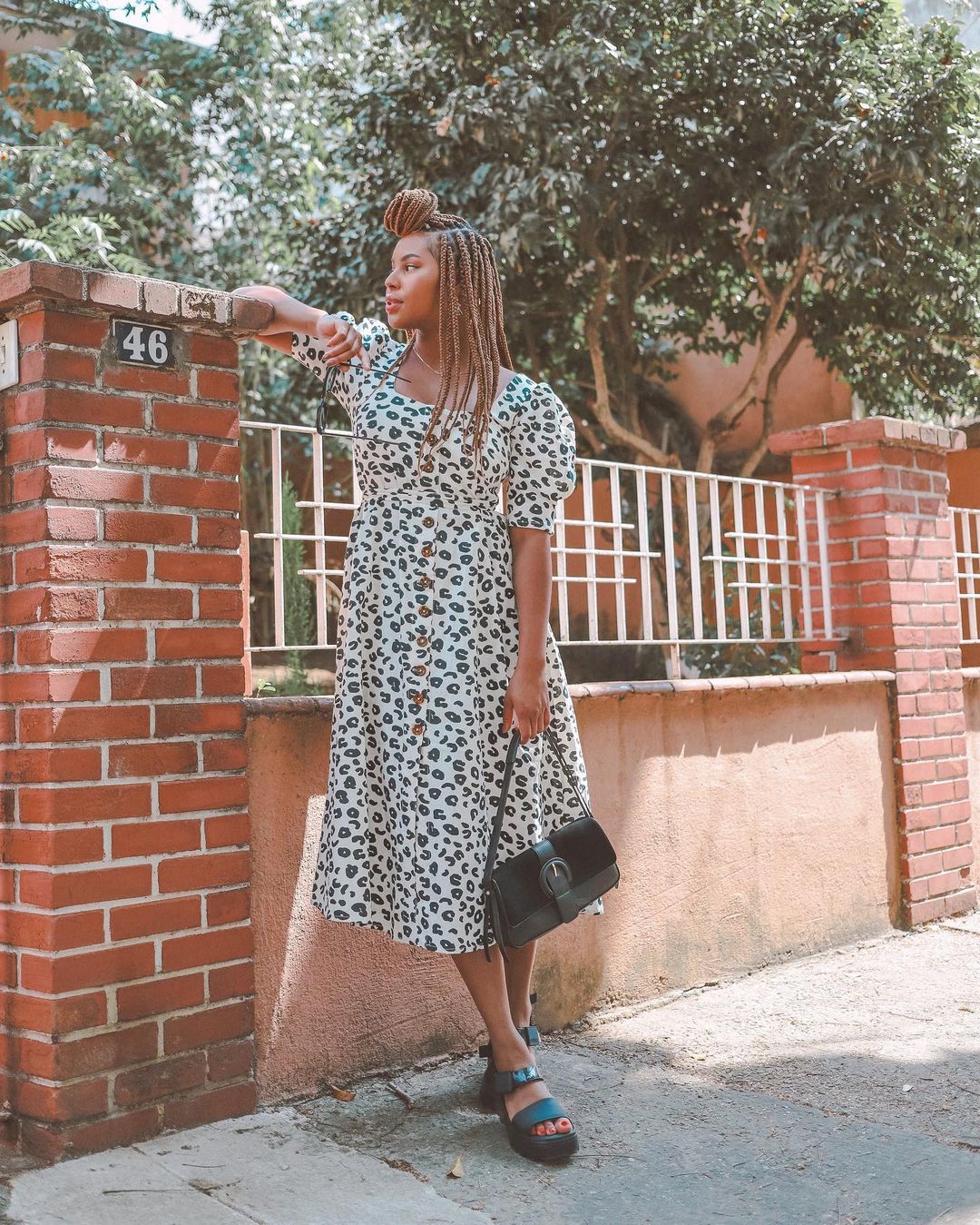 Garota usando vestido com estampa de oncinha preta e branca, segurando uma bolsa preta e usando uma sandália preta. Ela está olhando para o lado, com uma das mãos apoiadas em uma mureta de tijolinhos.