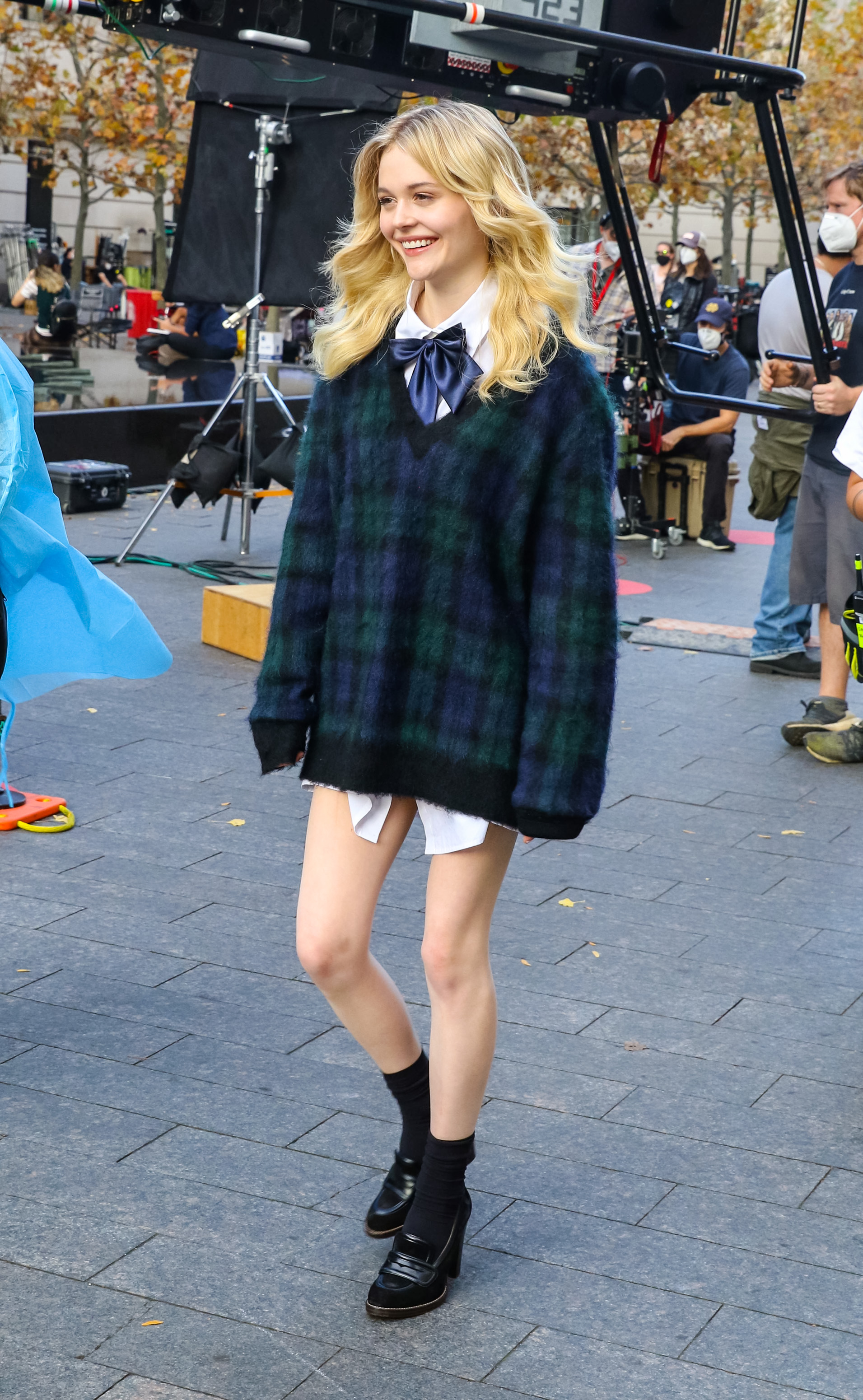 Gossip Girl Emily Alyn Lind nas gravações da nova Gossip Girl. Ela está usando um suéter xadrez por cima de uma camisa branca, um oxford preto de salto com meia preta. Ela está sorrindo enquanto anda e não olha para a câmera.