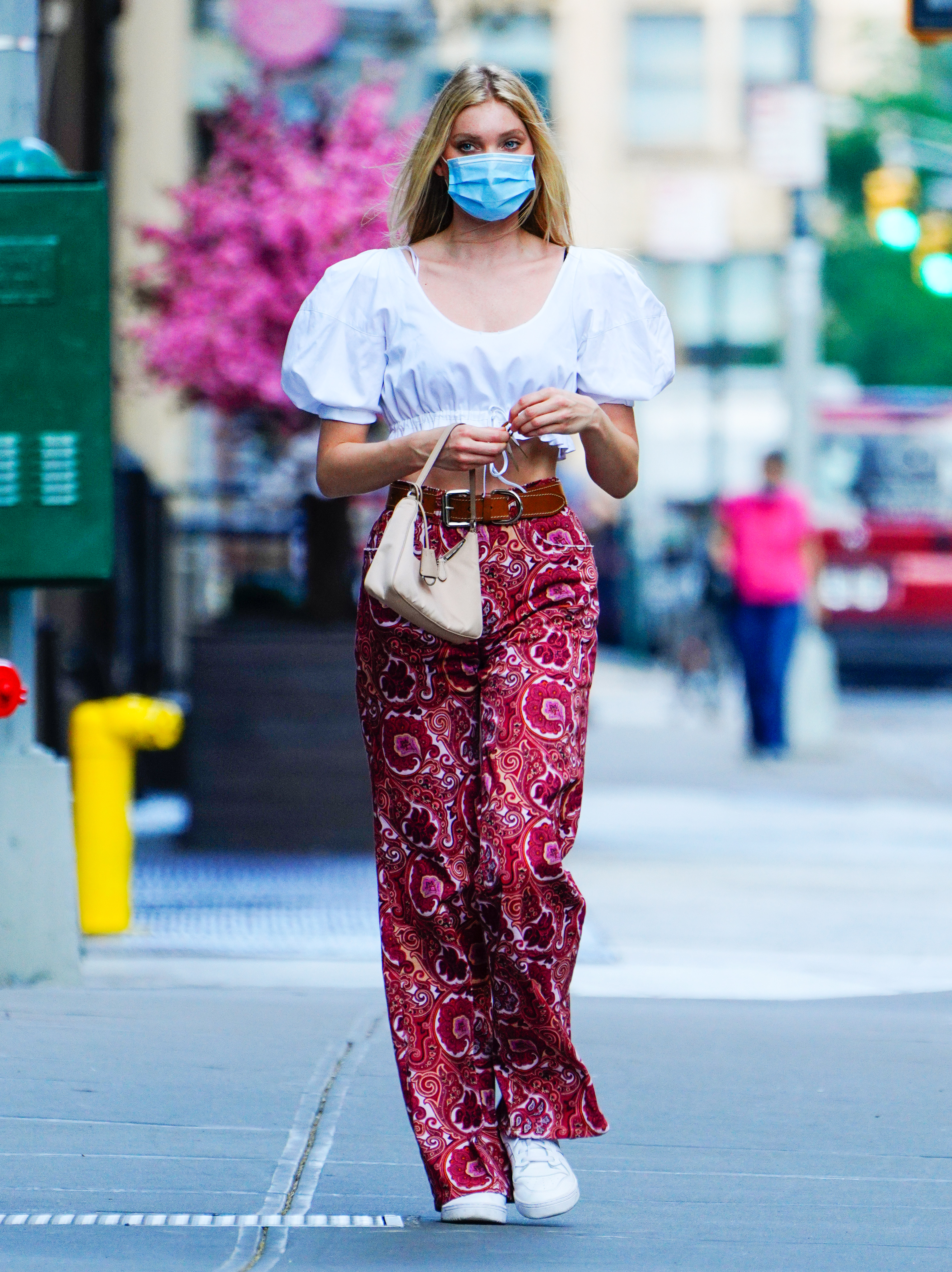 look-do-dia-elsa-hosk-boho-mangas-bufantes Elsa Hosk usando top cropped branco de mangas bufantes, calça vermelha com estampa paisley e tênis branco. Ela segura uma bolsinha rosa pastel em uma das bolsas e usa uma máscara de proteção azul e branca no rosto.