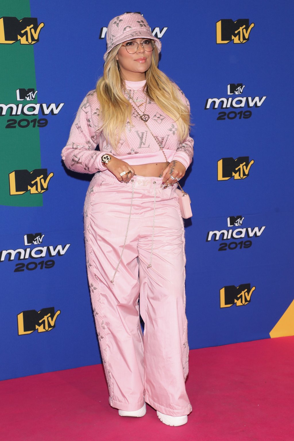 Karol G usando conjuntinho rosa formado por moletom cropped da Louis Vuitton e calça cargo, que é a cara dos anos 2000, além de um bucket hat rosa, bolsinha transversal e um tênis branco de plataforma