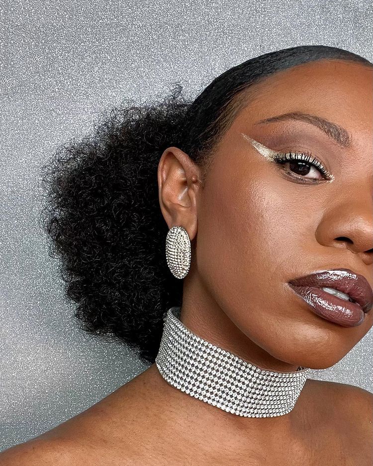maquiagem-festa-fim-de-ano-delineado-damatamakeup Metade do rosto de uma mulher, que está usando delineado dourado, gargantilha e brinco de brilhos e cabelo preso em um rabo baixo