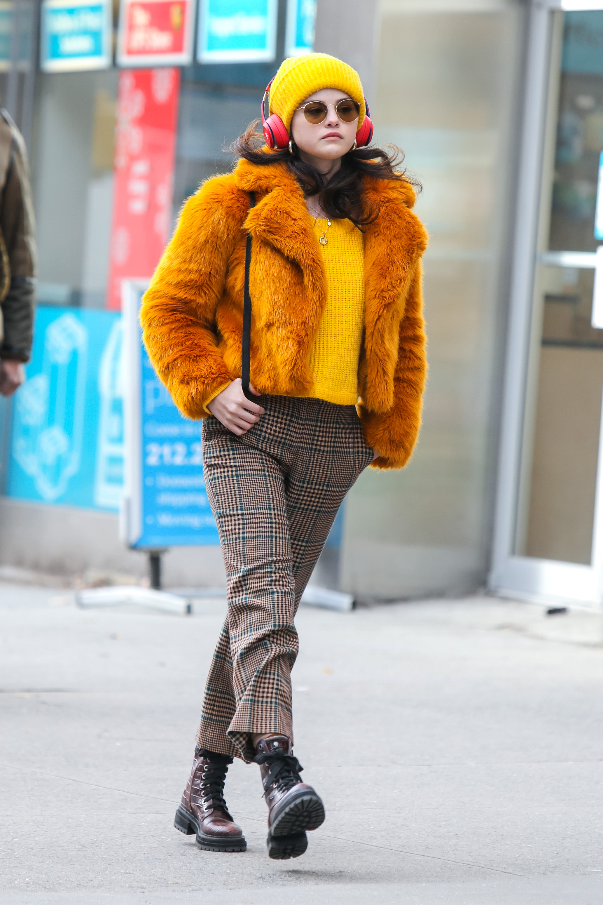 look-do-dia-selena-gomez-cor-pantone-2021 Selena Gomez usando suéter e gorro amarelos, casaco de pelos laranja, calça xadrez, coturno e óculos de sol