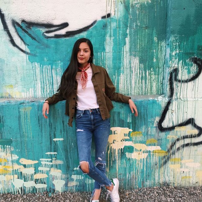 Looks Olivia Rodrigo Olivia Rodrigo usando calça jeans rasgata, camiseta branca, jaqueta verde militar e um lenço amarrado no pescoço. Ela está encostada em uma parede
