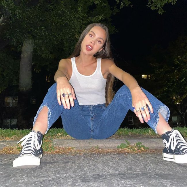Looks Olivia Rodrigo Olivia Rodrigo sentada usando calça jeans, blusa regata branca e tênis preto
