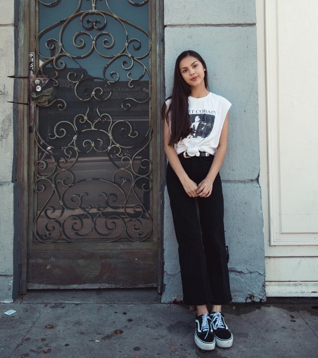 Looks Olivia Rodrigo Olivia Rodrigo usando calça e tênis pretos, camiseta branca com estampa preta e nó na região da barriga