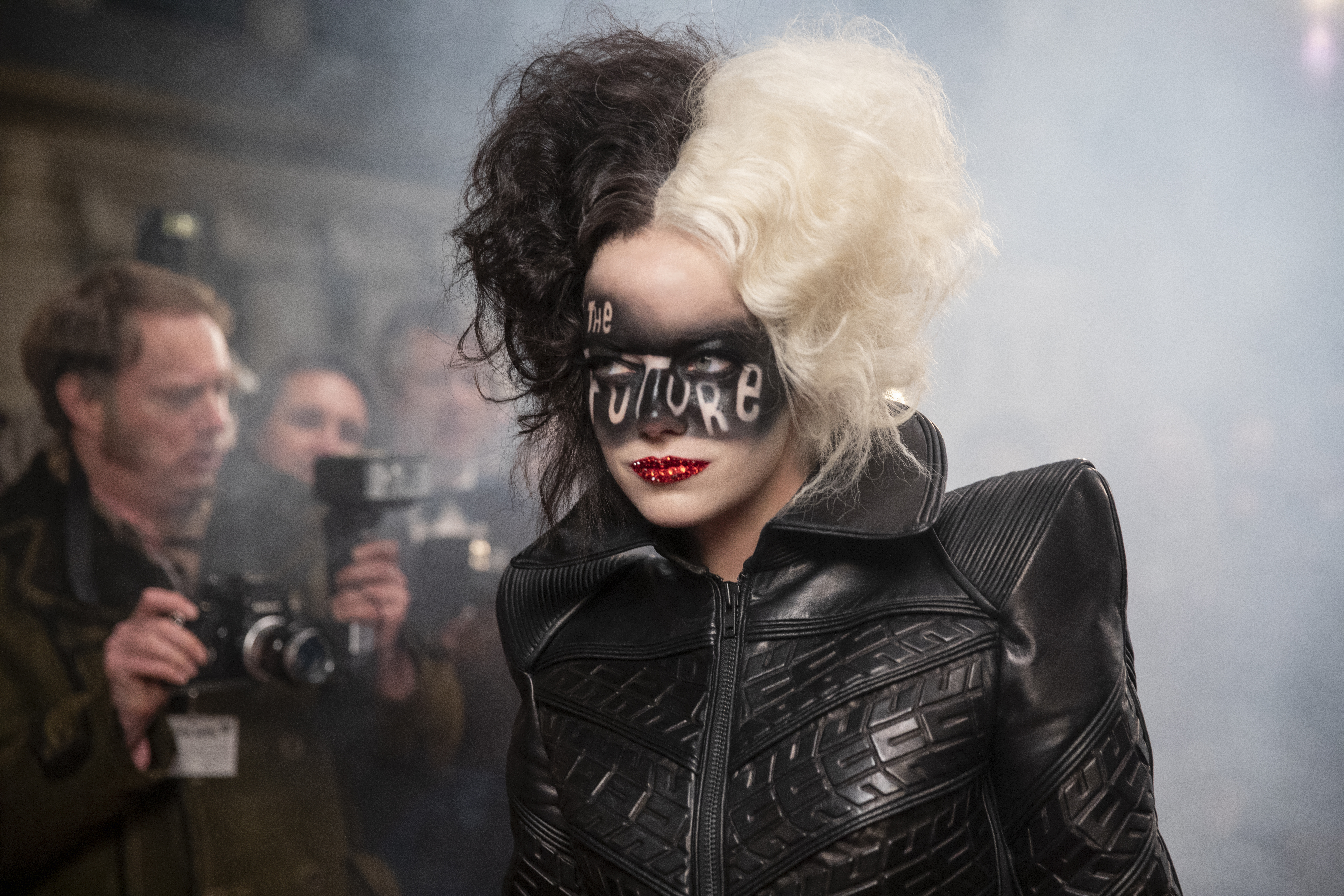 CRUELLA Emma Stone caracterizada como Cruella com metade do cabelo preto, outra metade loiro platinado com make escura que escreve