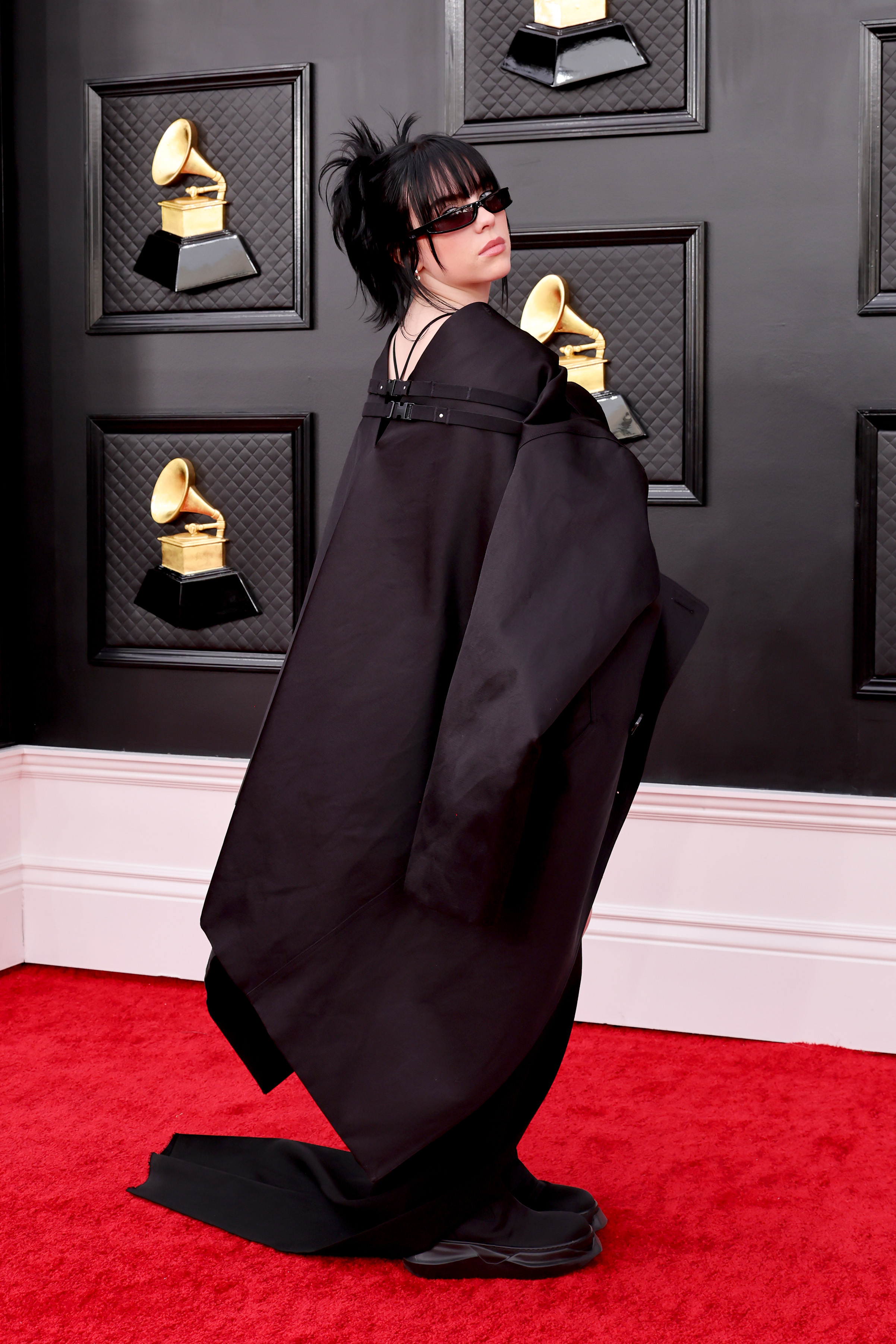 64th Annual GRAMMY Awards – Arrivals Imagem mostra Billie Eilish no tapete vermelho da 64ª edição do Grammy Awards usando um roupa-casulo preta. Ela está posando quase que costa e usa óculos escuro.