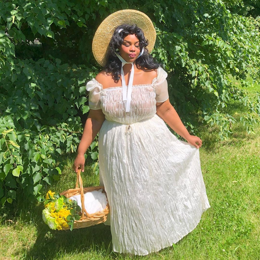 look-cottagecore-iridessence-02 garota no campo usa vestido branco com decote ciganinha a saia rodada, além de um chapéu com uma fita amarrada no queixo