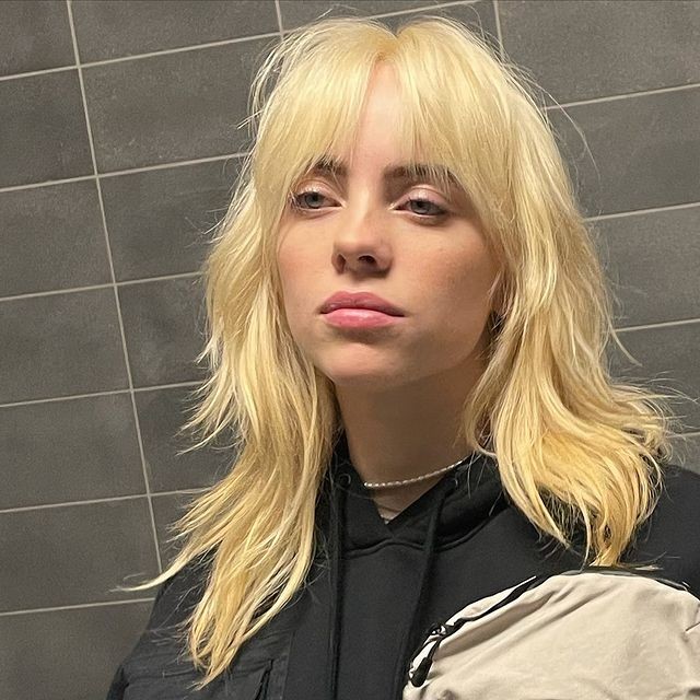 Cantora Billie Eilish com corte Shaggy Hair e expressão séria, encostada na parede, usando moletom preto, atrás dela podemos observar azulejos cinza.