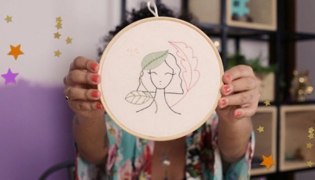 Mulher segura bastidor branco com desenho de um bordado de uma jovem com cabelo curto. Não conseguimos ver o rosto de quem segura o bordado, mas, conseguimos ver suas unhas pintadas de vermelho e sua camisa com detalhes florais.