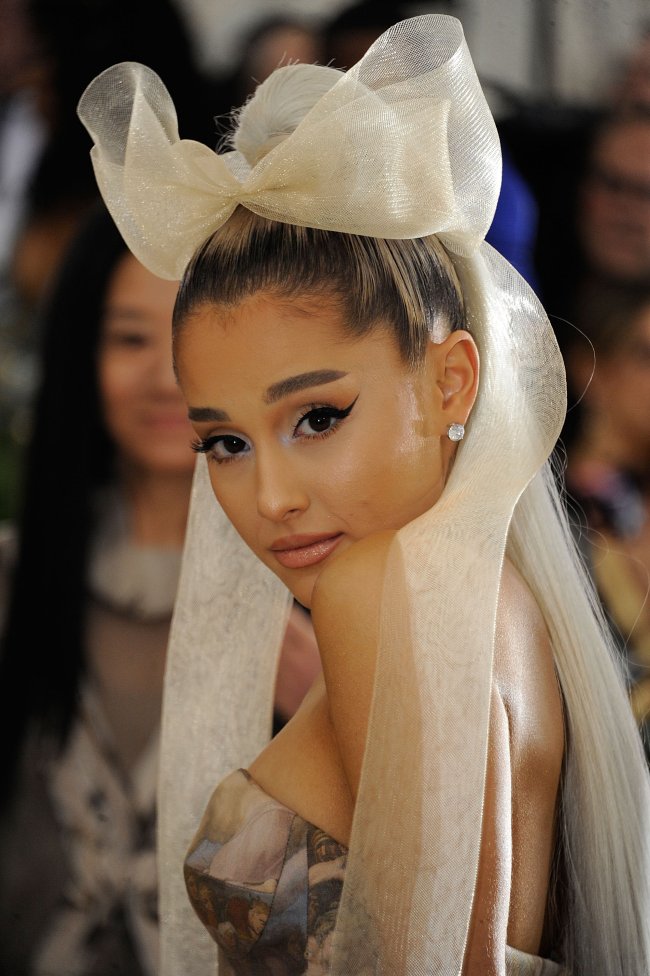 Ariana Grande Ariana Grande posando para foto virada de lado, com expressão séria, com a cabeça levemente abaixada, com grande laço na cabeça e vestido cinza tomara que caia.