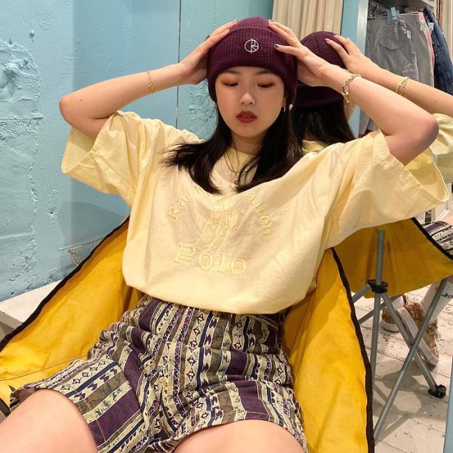Hina do Now United Hina, do grupo Now United, usando toca vermelha bordô, camiseta larga, short cintura alta, sentada em uma cadeira amarela e posando com as mãos na cabeça.