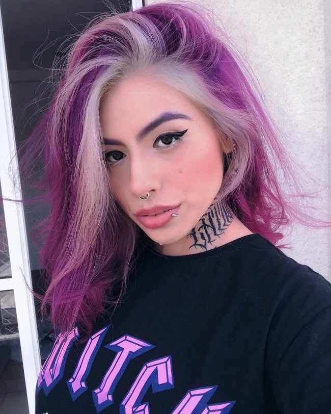 Jovem com cabelo roxo e platinado, de lado, olhando para a câmera, nos olhos delineado preto, no pescoço uma tatuagem que não conseguimos ler o que esta escrito pois na foto só aparece uma parte, camiseta preta, com witch escrito de roxo.