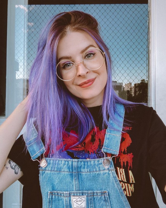 Jovem com uma das mãos na cabeça, com o cabelo roxo, usando óculos, com camiseta preta e jardineira jeans.