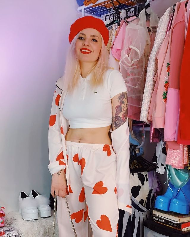 Jovem posa para foto com conjunto branco com corações rosa, conjunto composto por camisa de botões aberta e calça. Além disso, ela usa uma camiseta cropped branca, boina vermelha, e batom vermelho. Ela posa em frente a sua arara de roupas com as mais variadas peças. Sua expressão é de felicidade.