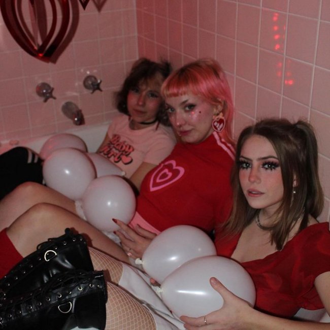 Looks inspirados na aesthetic lovecore Três jovens encaram a camera, sendo duas com uma expressão séria, todas seguram bexigas brancas, a primeira da esquerda para direta usa camiseta rosa clara, a segunda camiseta vermelha com coração no centro, e a terceira blusa vermelha com decote e podemos ver sua bota preta. Elas estão ao que parede sentadas dentro de uma banheira, podemos ver os azulejos do banheiro na foto.