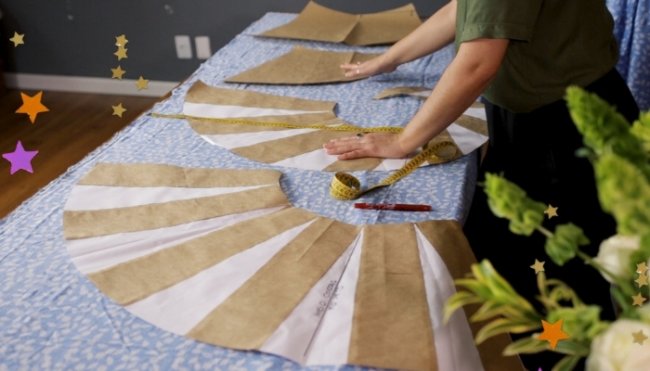 Mesa com vários moldes de roupas, podemos ver mãos trabalhando nos moldes, fita métrica, canetas e lápis espalhados pela mesa.