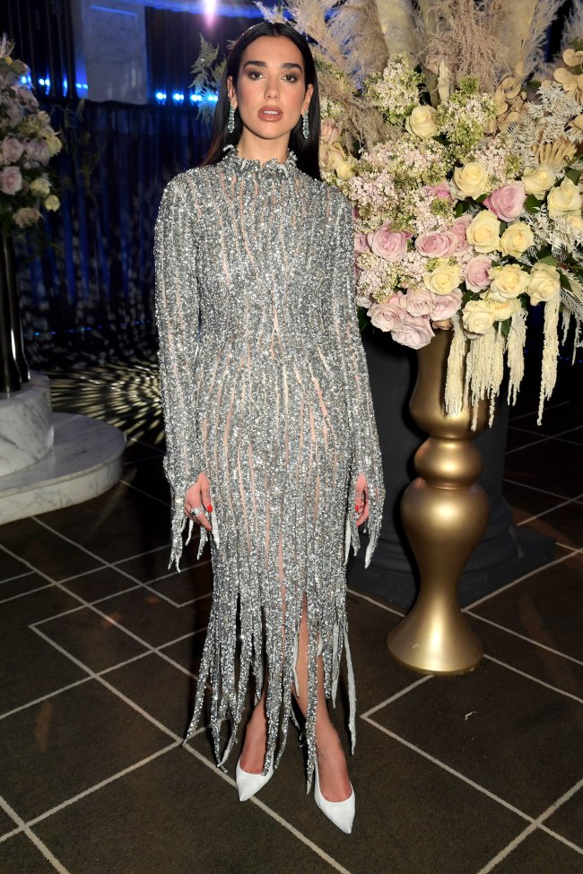 29th Annual Elton John AIDS Foundation Academy Awards Viewing Party Dua Lipa posando para foto ao lado de um vaso dourado com flores brancas e cor-de-rosa, usando vestido prata com franjas e levemente transparente, com os dois braços parados ao lado de seu corpo encarando a câmera com uma expressão séria.