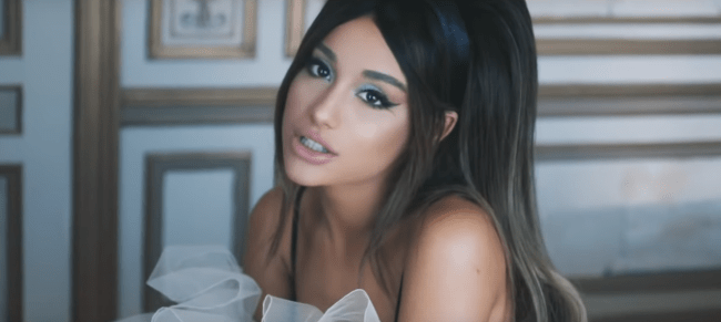 ariana-grande-boyfriend Ariana Grande sentada no chão, com expressão séria, com a cabeça levemente inclinada para cima, ao fundo da imagem podemos ver uma parede branca com detalhes em dourado. Print do clipe da música Boyfriend.