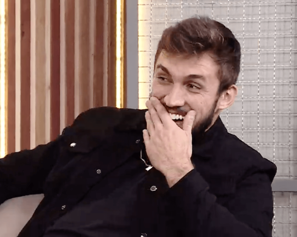 Arthur sentado, olhando para o lado, usando jaqueta e camiseta pretas. Ele sorri e coloca uma das mãos sobre a boc
