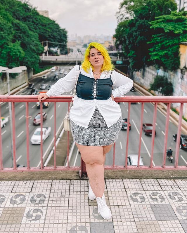 Garota usando camisa branca com corset preto por cima, além de saia xadrez preta e branca e coturno branco. Ela está com expressão facial séria, uma mão na cintura, e a outra segurando um corrimão. Ela está em uma ponte e, ao fundo, dá para ver uma avenida com carros.